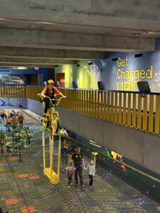 طفل يركب دراجة صفراء معلقة على سلك عالٍ فوق ممر المتحف، بينما يمشي الزوار أسفلها عبر منطقة مغطاة بشبكة. تزين المكان أضواء ملونة وشاشة عرض جدارية بعنوان "Get Charged Up!" (احصل على الطاقة!) ذات طابع علمي.